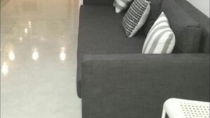Ein modernes Wohnzimmer mit einem dunkelgrauen Sofa, zwei gestreiften Kissen und einem weißen Sessel. Im Hintergrund ist eine Küche mit einer Steinrückwand sowie ein Essbereich mit einem weißen Tisch und Stühlen zu sehen.