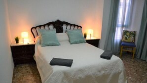 Un lit bien fait, recouvert d’une couvre-lit blanche et agrémenté de deux oreillers, avec deux tables de chevet surmontées de lampes, dans une pièce dont le sol est carrelé et où se trouve une chaise avec un coussin.
