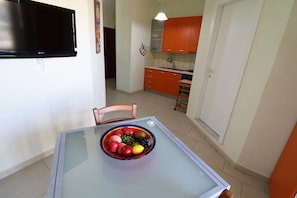 Uma área de refeições com mesa de vidro, uma tigela de frutas, uma televisão e armários cor de laranja ao fundo.