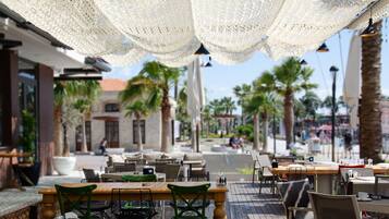 An outdoor cafe with tables and chairs, palm trees, and a building in the background.