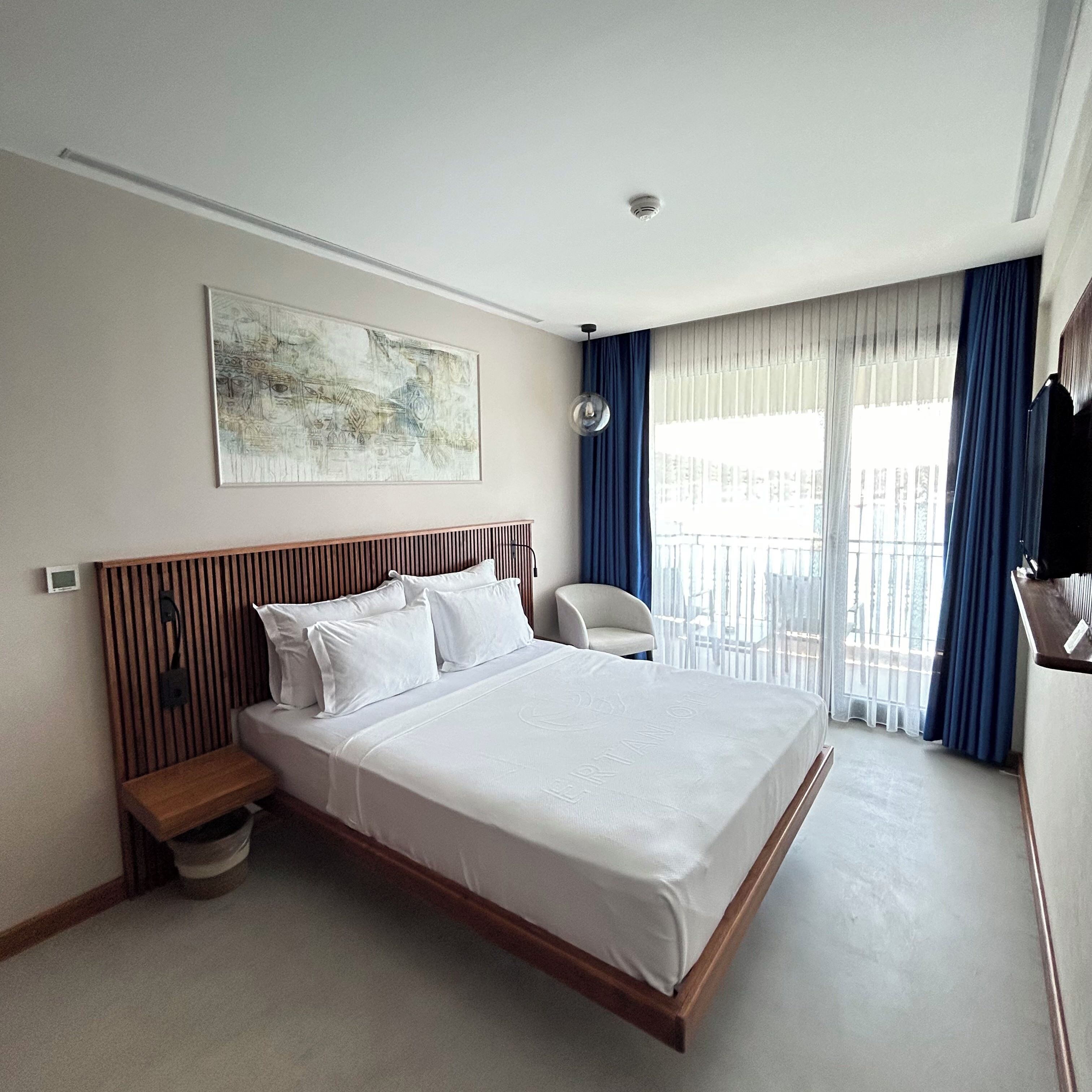 Une chambre d’hôtel avec un grand lit, une table de chevet, une chaise, une fenêtre avec des rideaux et un luminaire au plafond.