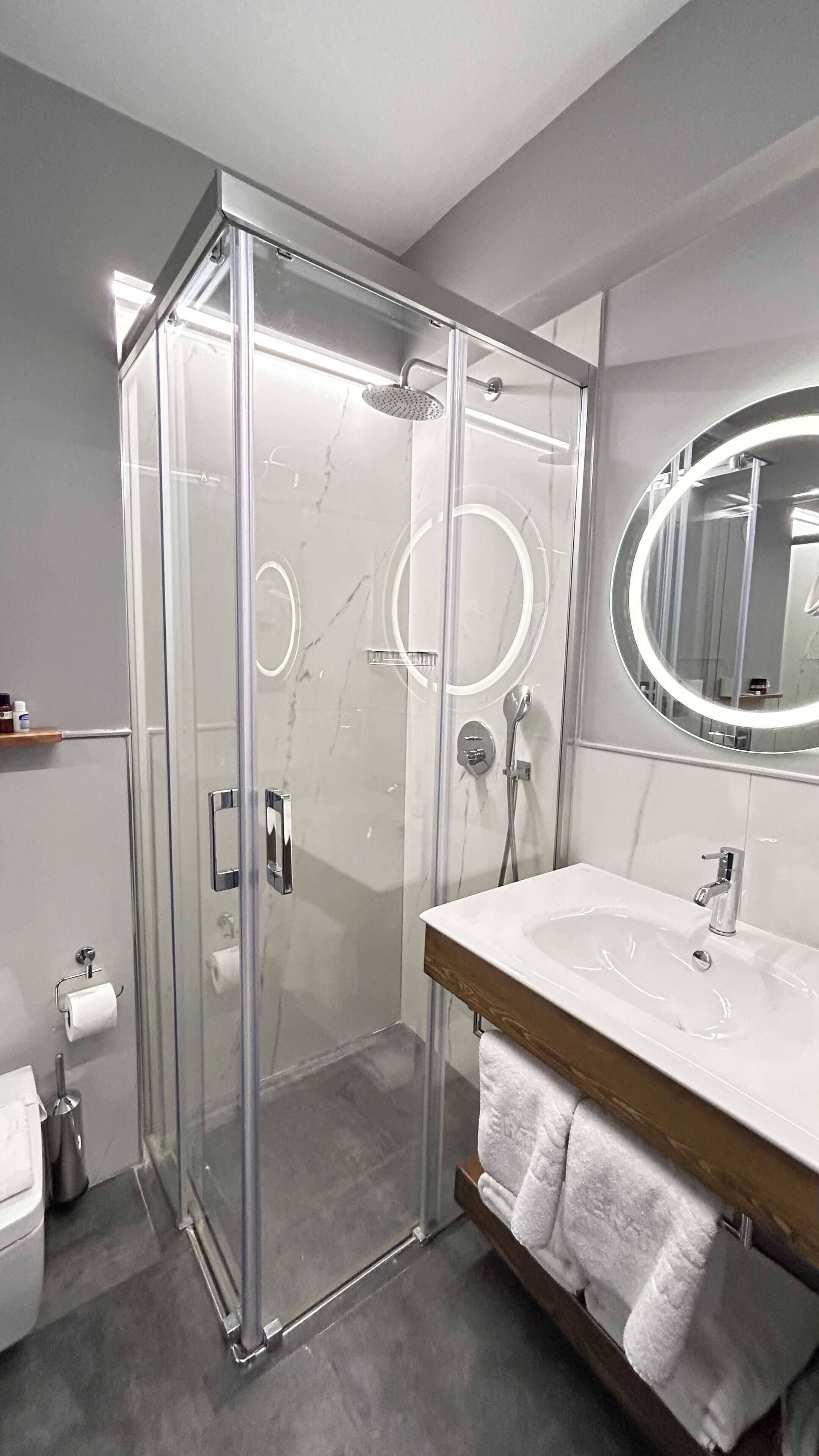 Un baño moderno con ducha envuelta en vidrio, un espejo redondo y un mueble de tocador de madera con lavamanos.
