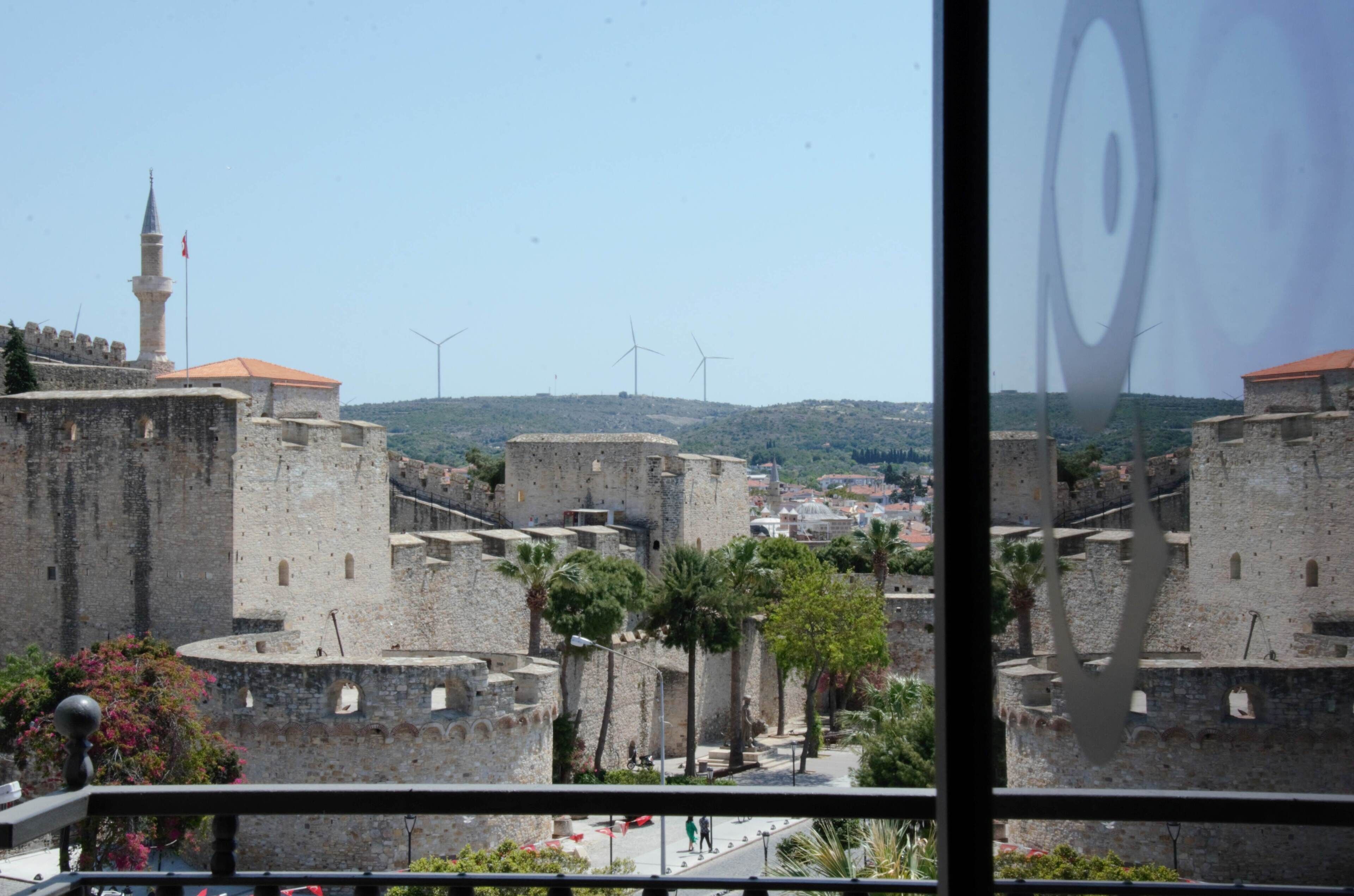 Une forteresse historique en pierre, dotée d’un minaret imposant, entourée de verdure et avec, en arrière-plan, des éoliennes modernes.