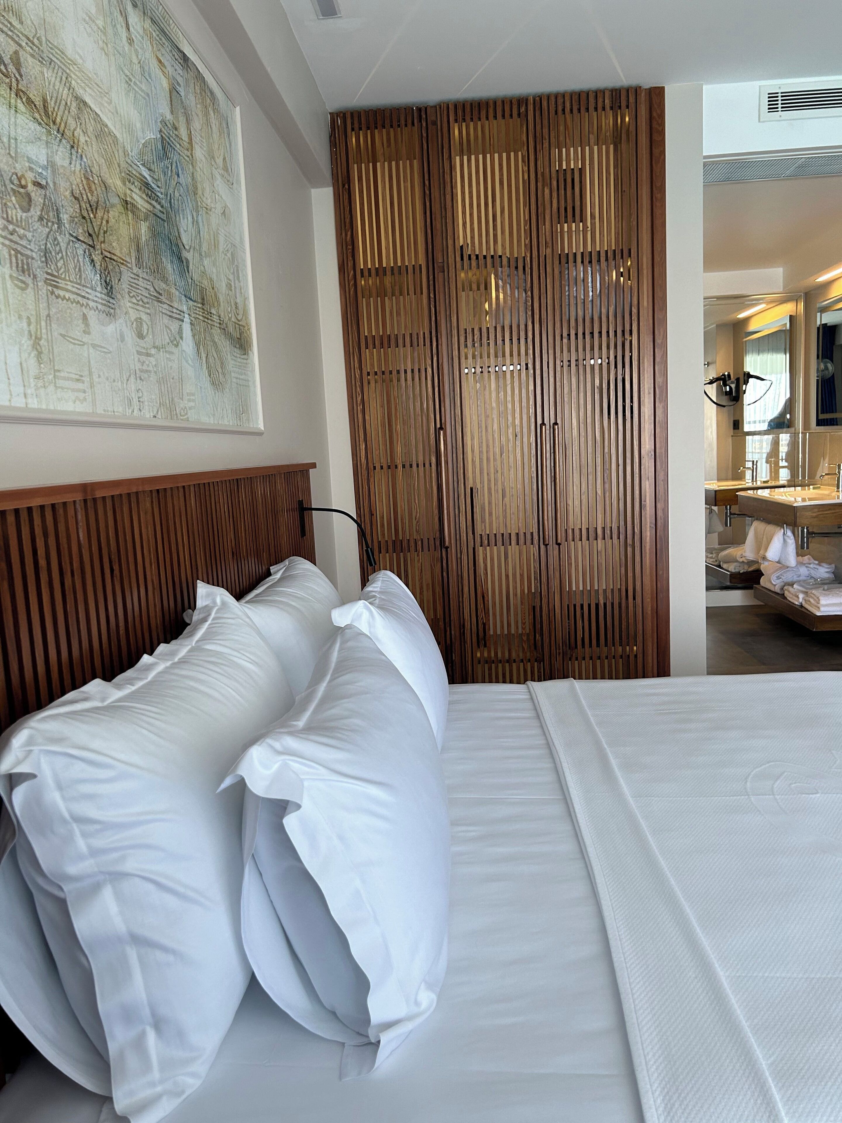 Habitación de hotel con cama, almohadas, separador de madera y una amplia pintura abstracta, con un baño visible a través de una puerta abierta.