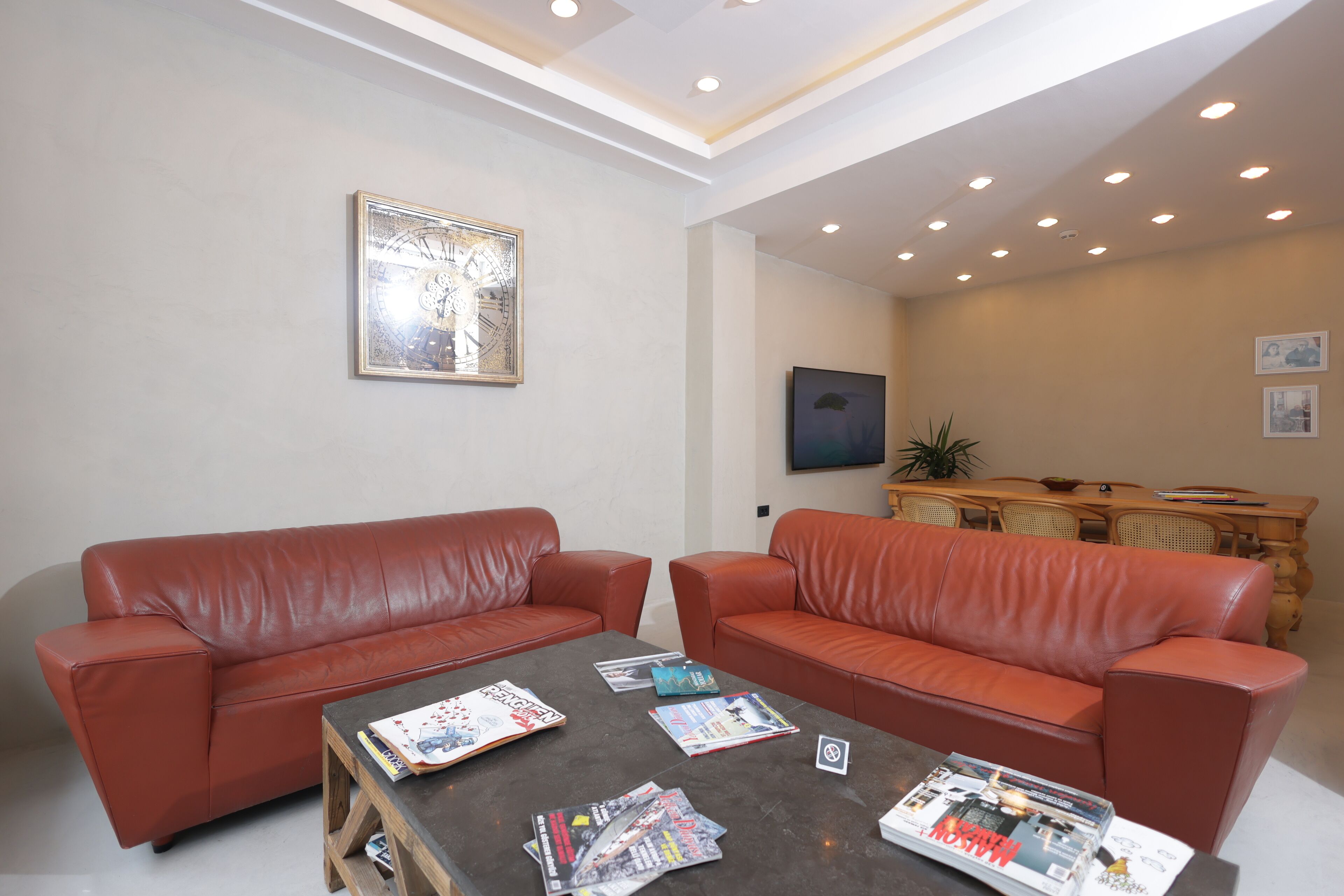 Una sala moderna con dos sofás de cuero rojo, una mesa de centro con revistas, un televisor de pantalla plana y un comedor con mesa y sillas de madera.