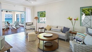 A spacious living room with a wooden floor, a round coffee table, a sofa, and a chair. There is a view of the ocean through large windows. - Southwinds - by ZenBreak (Mullins)