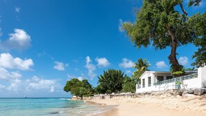 A beach with clear turquoise water, a white building with a balcony, and a large tree. - Southwinds - by ZenBreak (Mullins)