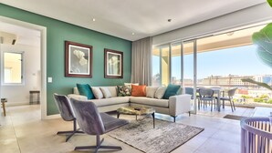 Un salon moderne avec un canapé gris, un fauteuil noir et une table basse. Deux tableaux encadrés ornent le mur, une grande fenêtre offre une vue sur les bâtiments et un balcon est aménagé avec du mobilier d’extérieur.