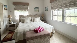 Une chambre à coucher comprenant un lit, des tables de chevet, une fenêtre ornée de barreaux en fer décoratifs et un ventilateur de plafond.