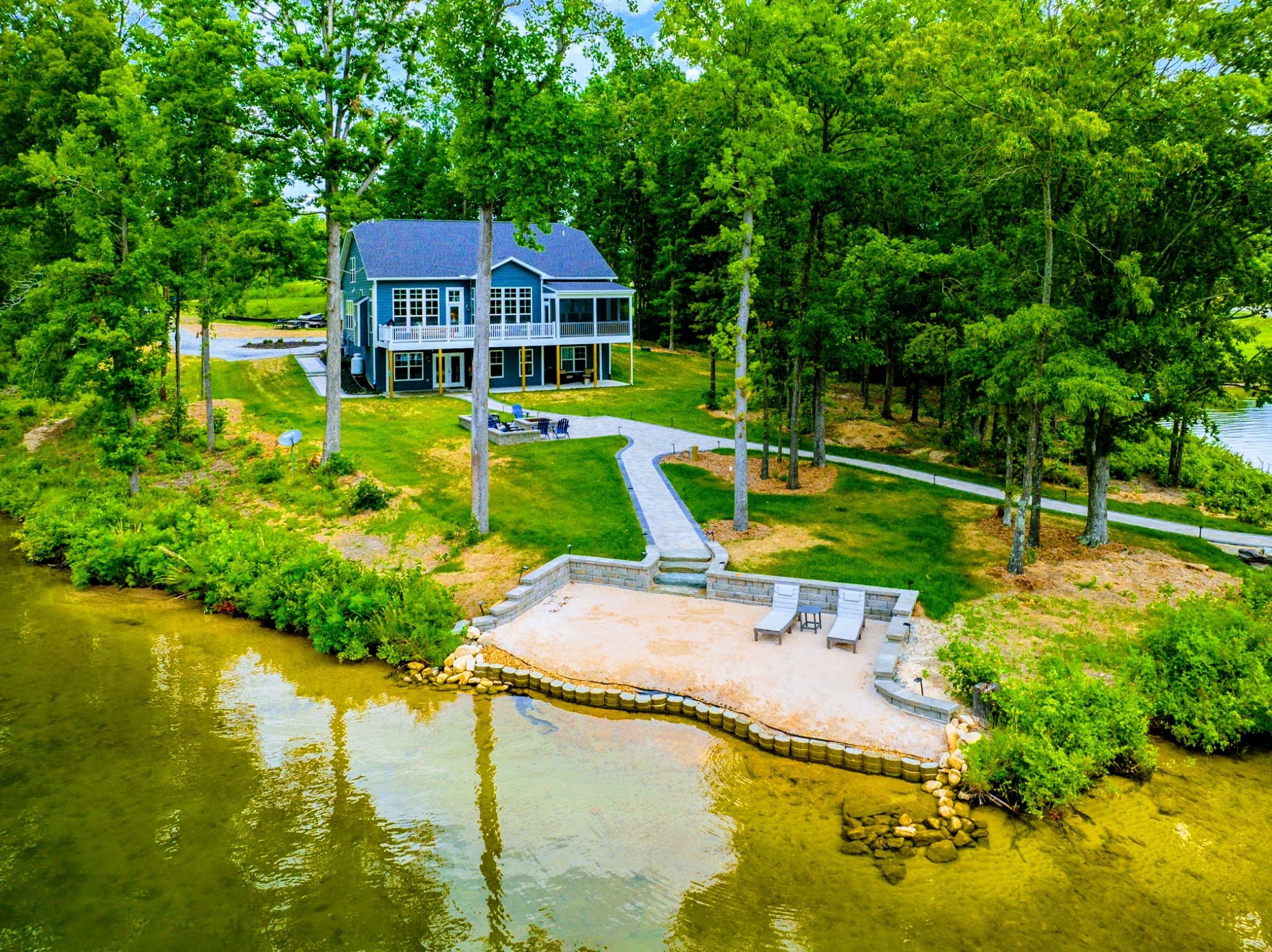A two-story house with a dock extending into a lake, surrounded by lush greenery and trees.