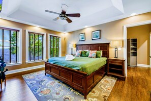 Un dormitorio con una cama de madera, ventilador de techo, mesita de noche, lámpara y vistas a la vegetación a través de las ventanas.