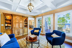 A spacious living room with a wooden cabinet, blue armchairs, a round coffee table, and large windows offering a view of the outdoors. - Huge 8BR Lakefront Lake Anna, Beach, Dock, Hot Tub (Bumpass)