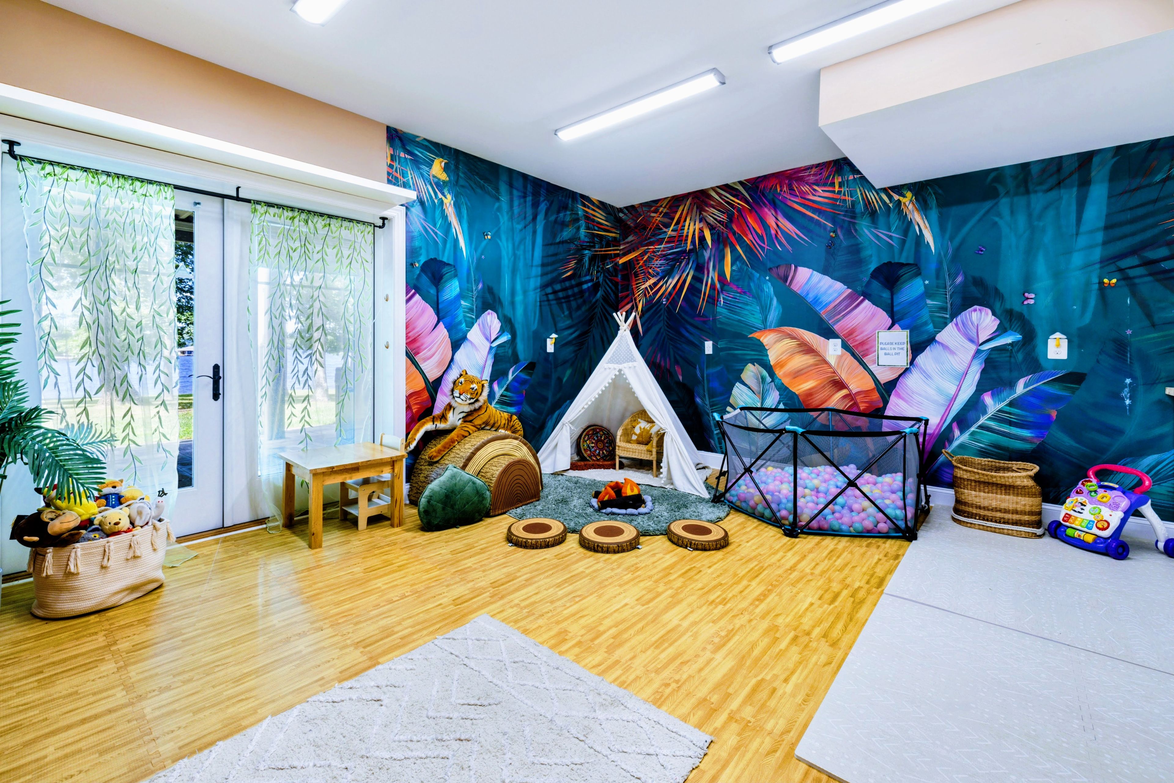 A room with a wooden floor, a white table, a teepee, a ball pit, and a wall with a tropical leaf mural.