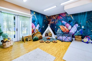 Una habitación con piso de madera, una mesa blanca, un tipi, una piscina de pelotas y una pared con un mural de hojas tropicales.
