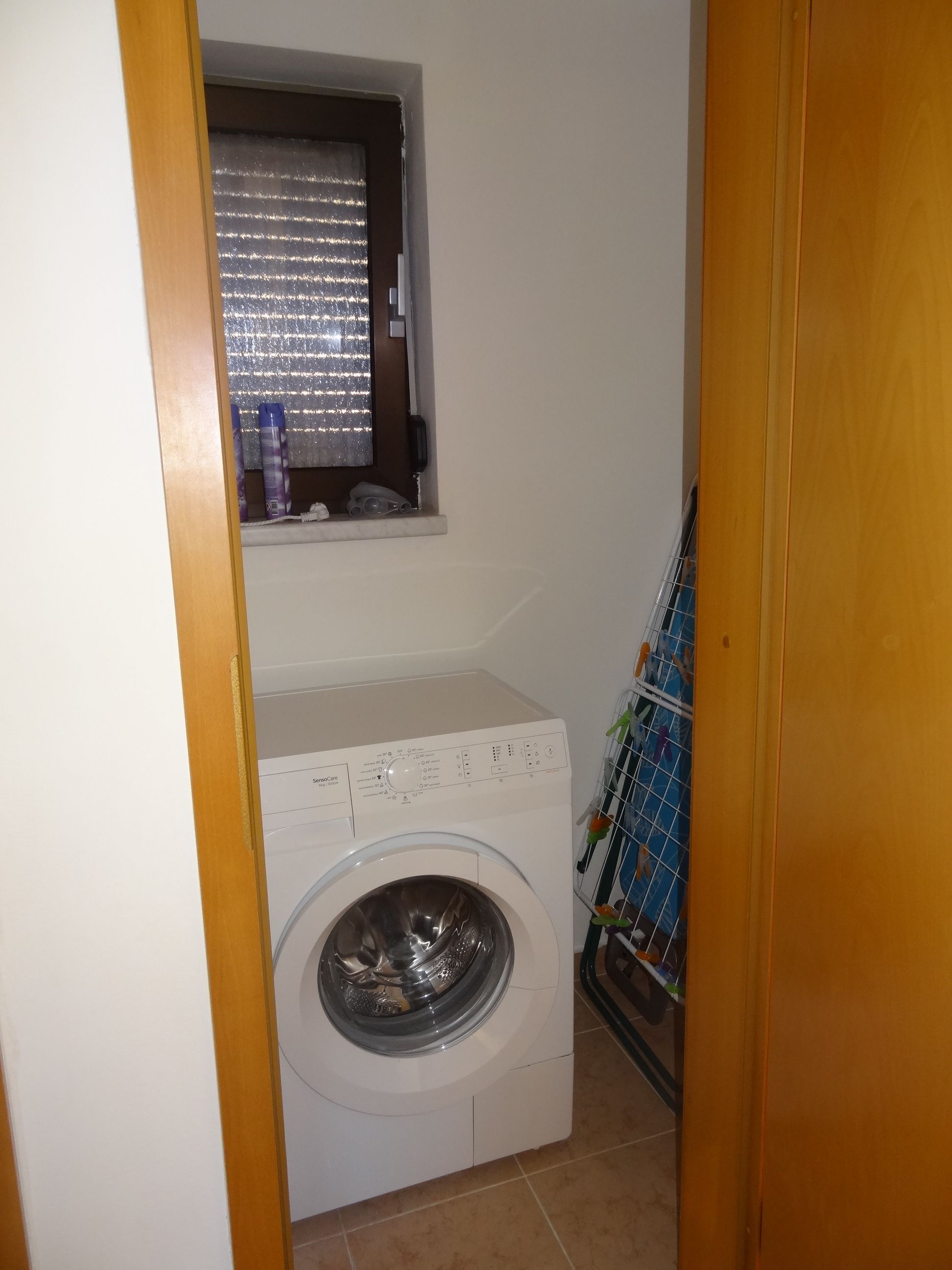 Un lavadero pequeño y funcional con lavadora de carga frontal, una ventana con persianas y un tendedero con ropa.
