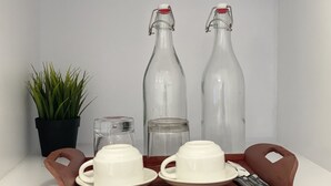 Una bandeja con dos tazas y platillos blancos, dos botellas de vidrio con tapas metálicas, un frasco pequeño de vidrio y una planta en maceta.