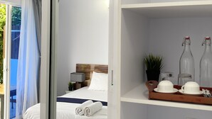 Habitación de hotel con cama, mesita de noche, lámpara, planta en maceta y estante con botellas y vasos.