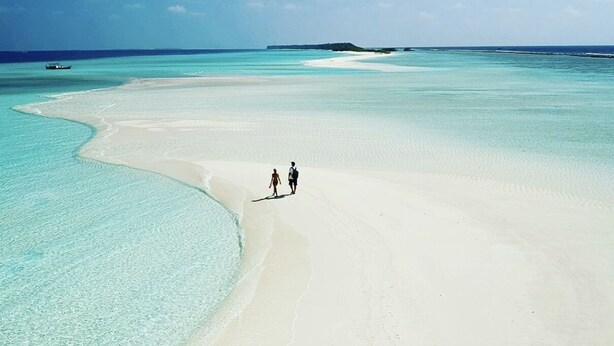 Una playa con dos personas caminando sobre la arena, un barco en el agua y una isla distante.