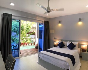 A modern hotel room with a large bed, a ceiling fan, and a sliding glass door leading to a balcony with potted plants. - Tides Dhigurah (Dhigurah)