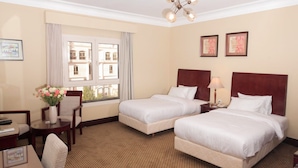 Quarto de hotel com duas camas, uma escrivaninha, uma cadeira, um vaso com flores e uma janela com vista para edifícios.