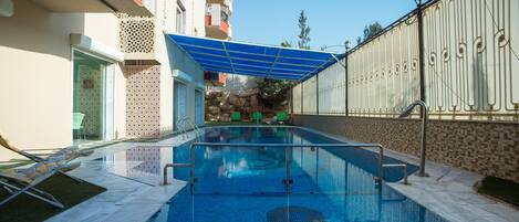 Una piscina con toldo azul, sillas verdes y un muro de piedra al fondo.