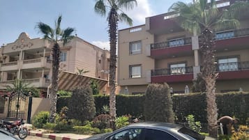 Un immeuble à plusieurs étages doté de balcons, de palmiers et d’une voiture garée devant.
