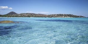 A coastal view with clear turquoise waters, a rocky outcrop, and a hill with buildings in the background.