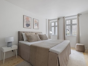 Un dormitorio con cama, mesita de noche, lámpara, obra de arte y una ventana con cortinas.