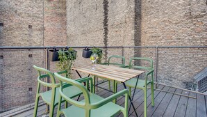 Une table en bois, entourée de quatre chaises, sur un balcon dont le fond est constitué d’un mur en briques.