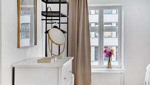 A room with a white dresser, a mirror, a black metal shelf, a beige curtain, and a window with a view of buildings. - Regent 7 - Large 3 Bdr Apt with Balcony in City Centre (København)
