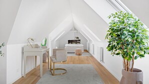 Un dormitorio estilo loft moderno con techo inclinado, un escritorio blanco con silla, una mesita de noche, cama con cubrecama acolchado y una planta grande en maceta.