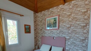 A bedroom with a stone-patterned wall, a bed with white bedding, a wooden ceiling, and a window with curtains. - Samos Island - Panorama Apartment with Jacuzzi (Koumeika)