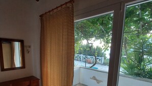Ein Zimmer mit Steinfußboden, einem Holztisch, einem Fenster mit Blick auf Grünflächen und einem karierten Vorhang.