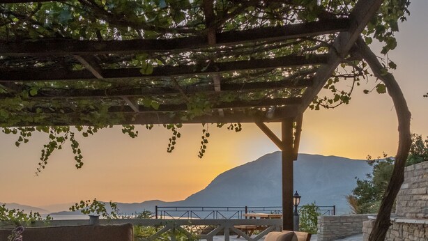 Un espacio al aire libre con mobiliario de madera y una pérgola, con vistas a un paisaje montañoso y un atardecer.