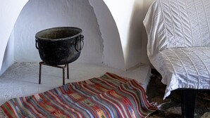 Una habitación con pared blanca, estante de madera con un cuenco azul, una olla negra sobre un soporte, una alfombra colorida y una cama con colcha blanca.