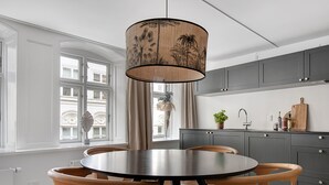 A modern kitchen with a round dining table, wooden chairs, a hanging light fixture, and a view of a building through the window. - Regent 4- Cute 1-Bdr Apt in Centre of Copenhagen (København)
