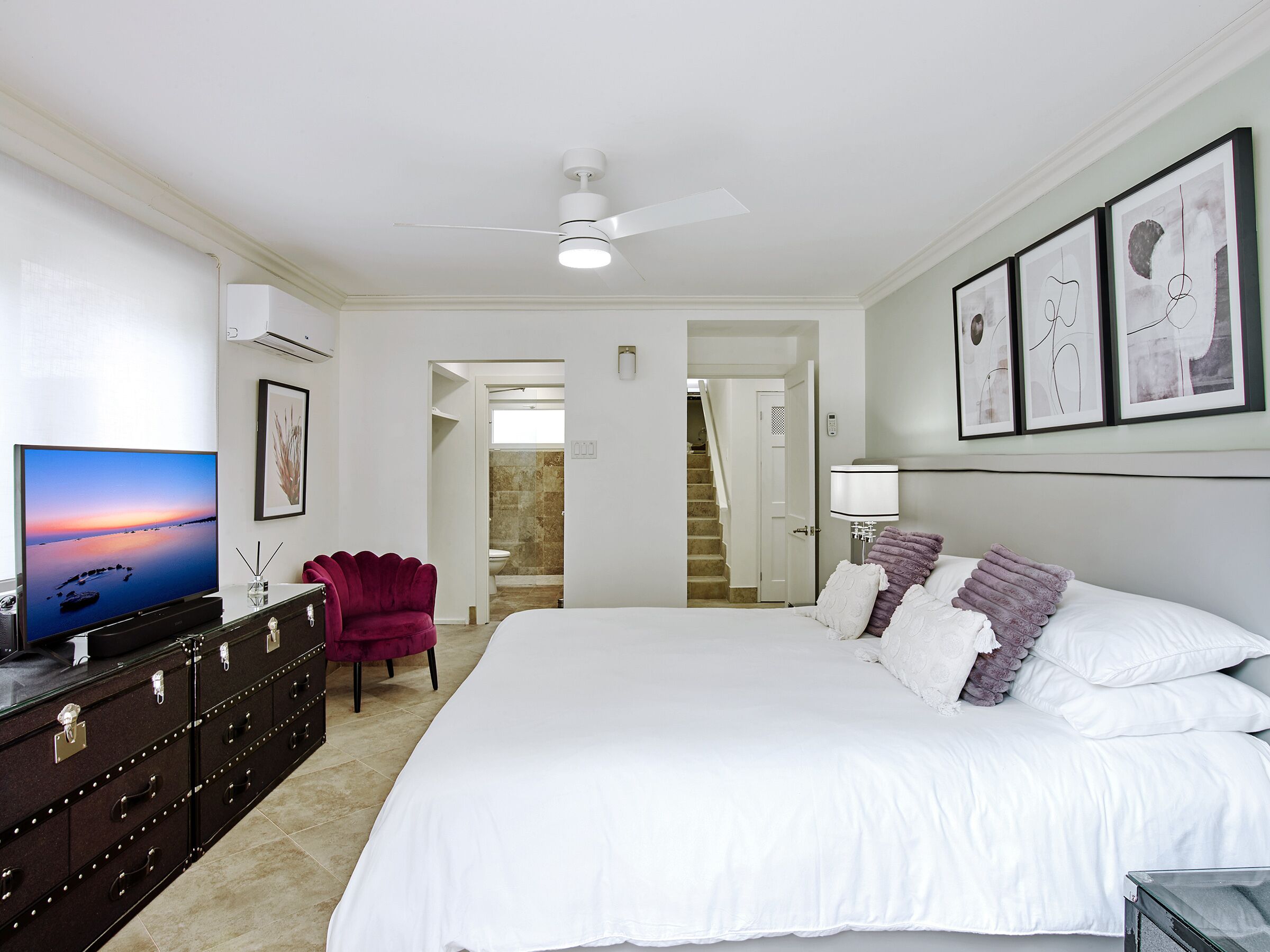 Een slaapkamer met een groot bed, een televisie op een dressoir, een plafondventilator en kunstwerken in een lijst aan de muur.