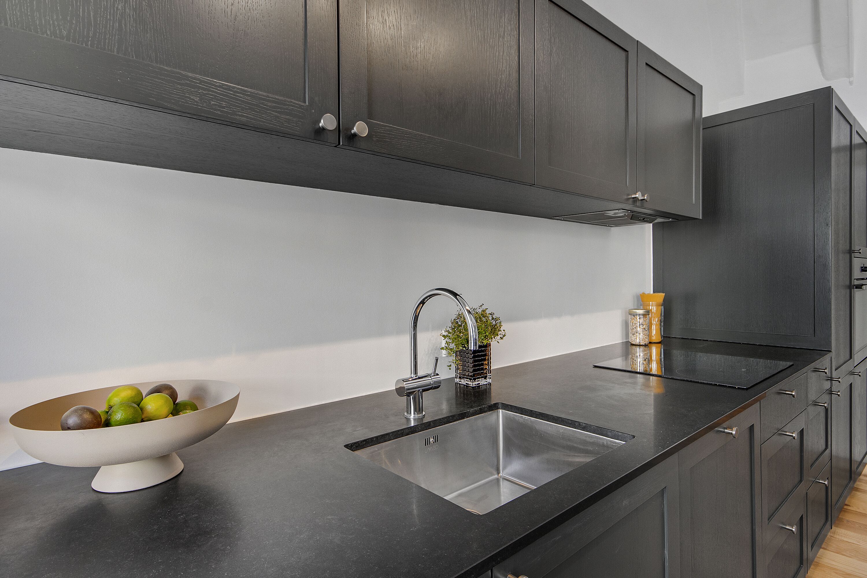 A modern kitchen with dark cabinets, a stainless steel sink, and a bowl of fruit on the counter.