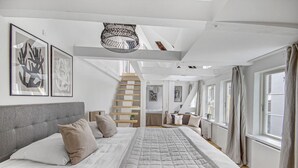 Ein geräumiges Schlafzimmer mit einem großen Bett, einer Treppe zu einer oberen Ebene und großen Fenstern mit Vorhängen.