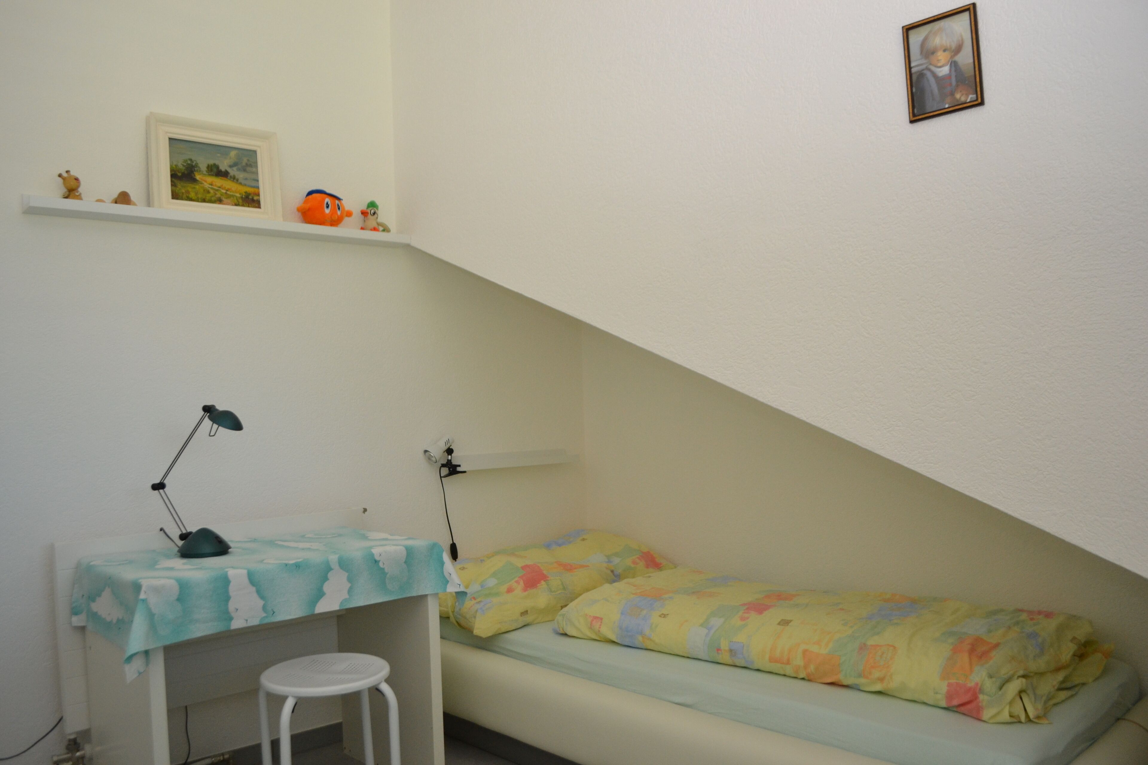 Um quarto pequeno com uma cama de solteiro, uma escrivaninha com cadeira, uma prateleira com brinquedos e um quadro na parede.