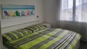 Et soveværelse med seng, et natbord, et vindue med gardiner og et maritimt maleri på væggen.