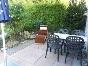 Un patio al aire libre con una mesa y sillas, rodeado de follaje y un seto.