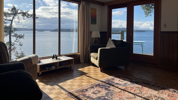 Habitación con ventanas amplias que ofrecen vista a un lago y árboles distantes. Hay un sofá, una mesita y una alfombra en el suelo.