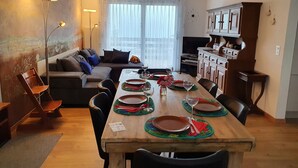 Un coin repas avec une table en bois dressée pour quatre personnes, entourée de chaises. On aperçoit un canapé et un téléviseur à l’arrière-plan.