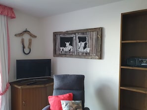 A living room with a TV, a chair with cushions, a wooden cabinet, and a wall-mounted artwork. - Studio for 2-4 guests with balcony and panorama view (Emmetten)