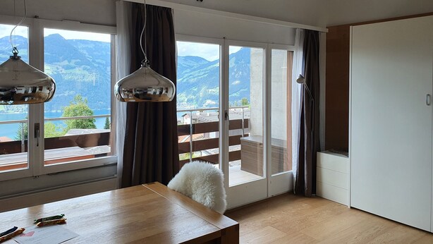 A room with a wooden table, a white chair, and a view of mountains through large windows.