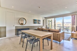 A modern kitchen with a dining table, chairs, a sofa, and a large window offering a view of buildings and greenery.