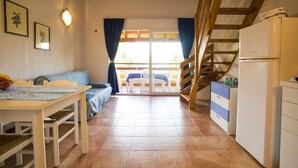 Une cuisine chaleureuse avec une table à manger, un canapé et un réfrigérateur. Un escalier mène à un balcon équipé d’une table et de chaises.