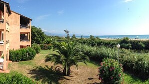 A multi-story building with balconies, a garden with palm trees, and a view of the beach.