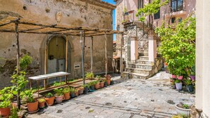 Eine enge Kopfsteinpflasterstraße mit einem rustikalen Gebäude auf der einen Seite und einer Treppe, die zu einem Balkon führt, auf der anderen. Übertöpfe mit Pflanzen und ein Holztisch stehen unter einer Pergola.